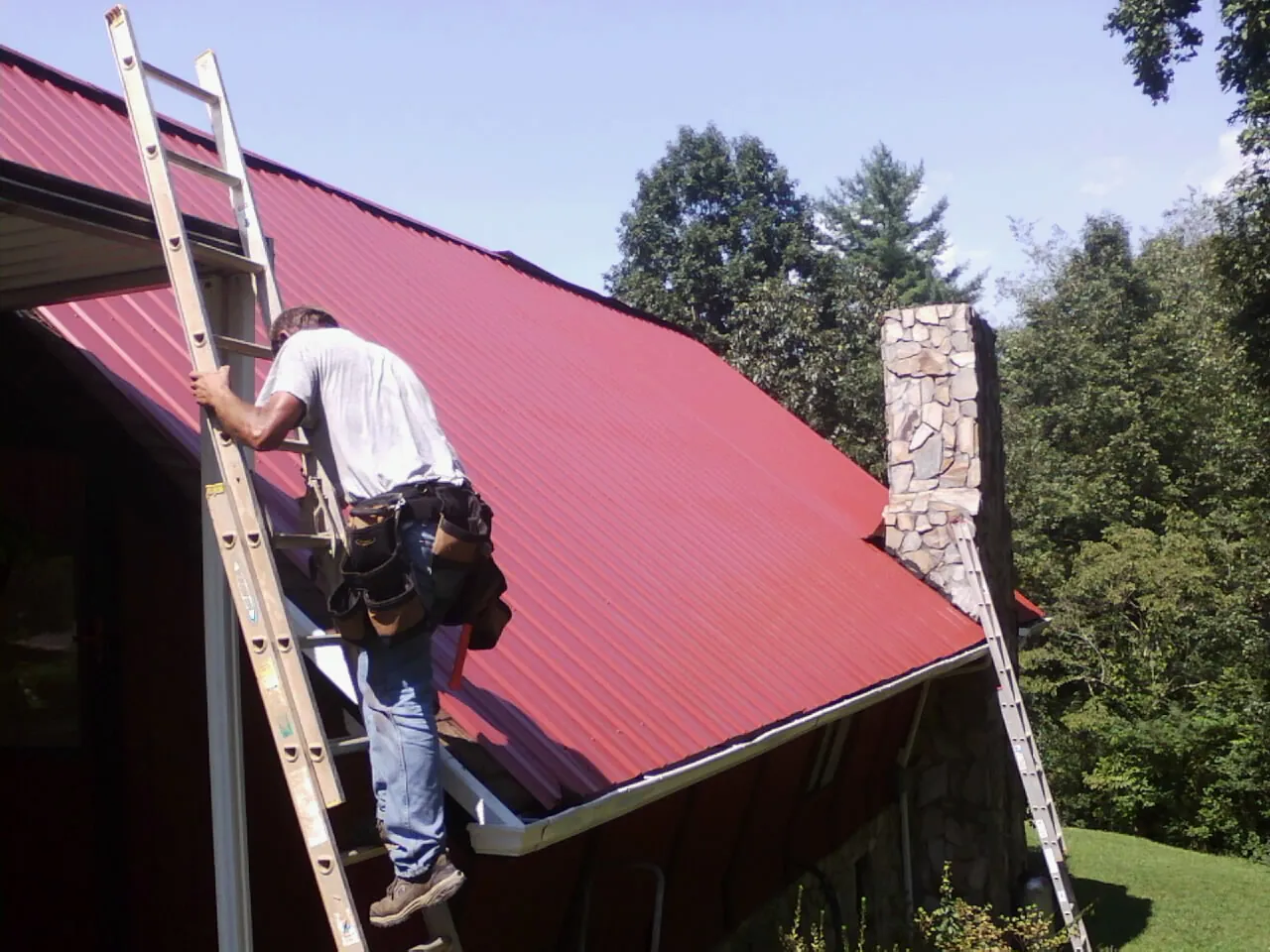 Roofer on a ladder beside a red metal roof and stone chimney — Soffit & Fascia Repair by Roofing Brothers in Sugar City